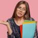 Confused puzzled brunette intelligent woman geek has clueless expression, clasps hand in bewilderment, holds colourful folders with important inforrmation, doesnt know what expect, stands indoor