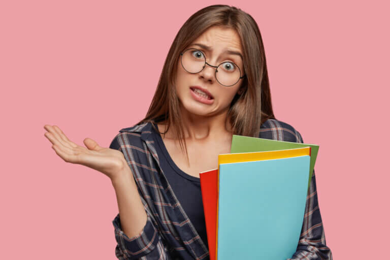 Confused puzzled brunette intelligent woman geek has clueless expression, clasps hand in bewilderment, holds colourful folders with important inforrmation, doesnt know what expect, stands indoor